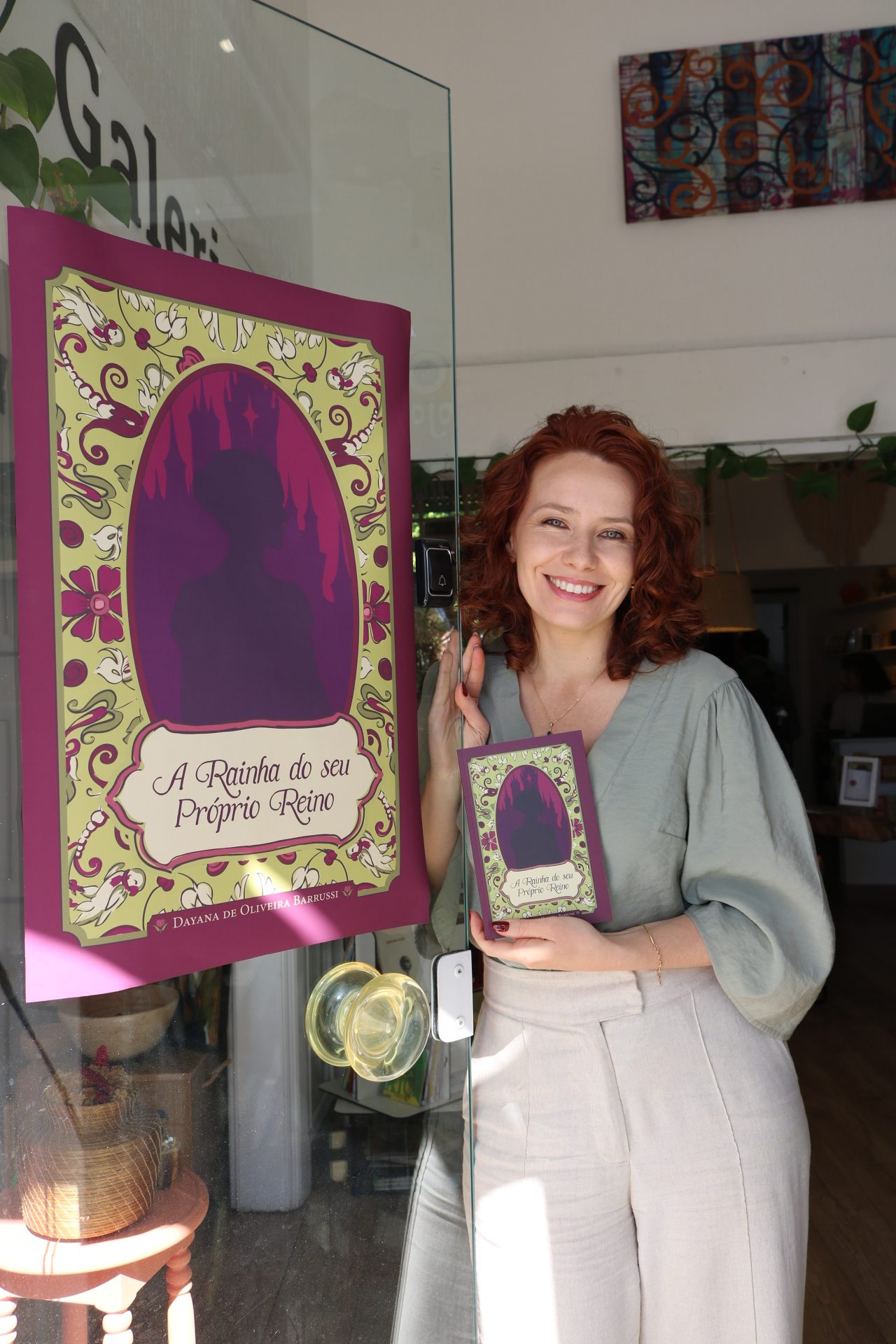 Dayana Barrussi com o livro ao lado do banner da Rainha
